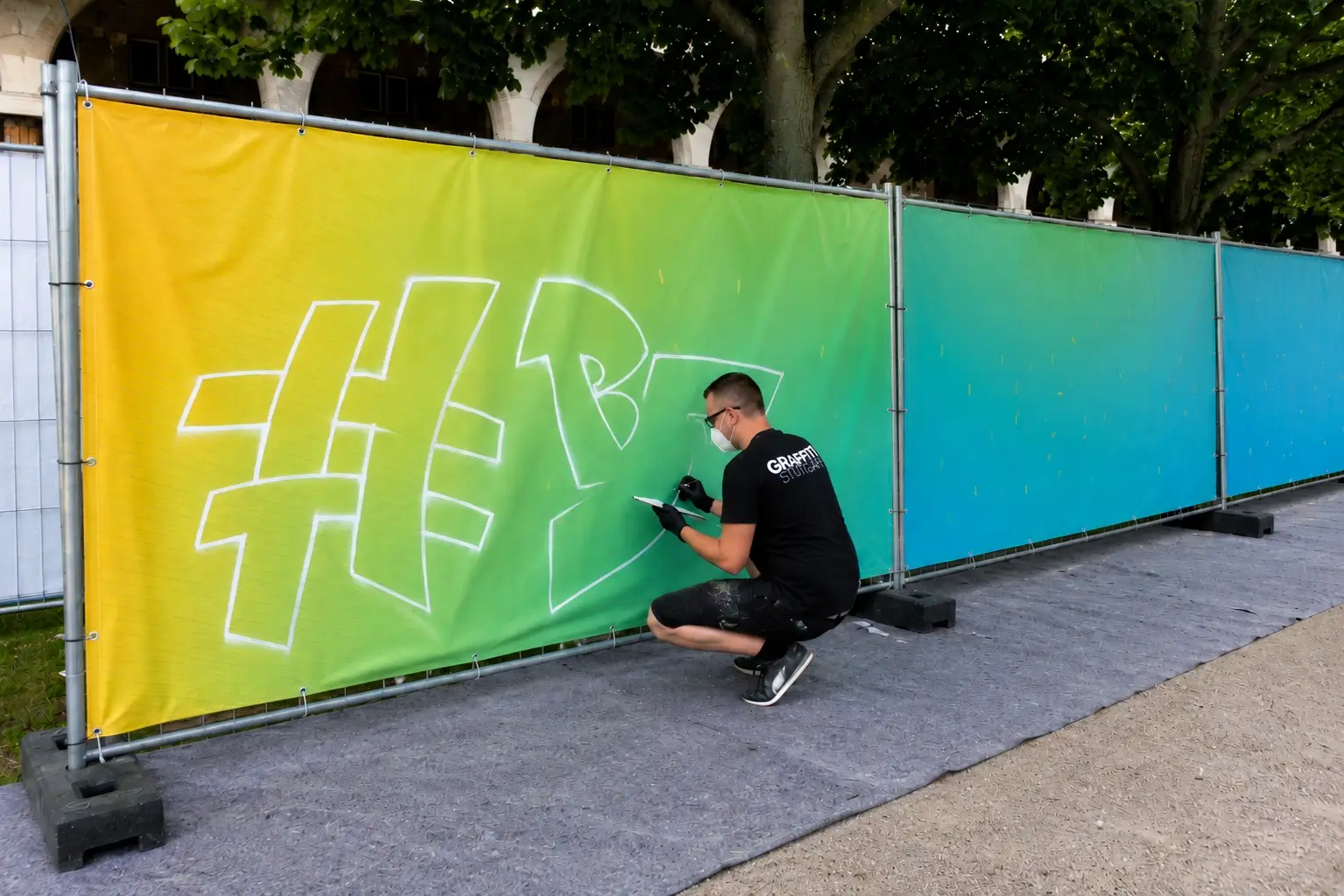 Mehrere Bauzäune mit farbigen PVC-Bannern, auf denen ein Künstler eine Graffiti-Skizze für eine Mitmachaktion vorbereitet.